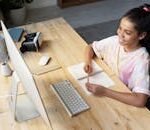 A young girl smiling while studying online at home, using a computer and taking notes.